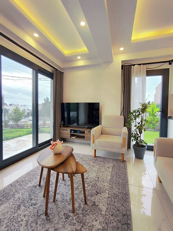 A modern living room with a large TV, comfortable seating, a coffee table, and a view through large windows.
