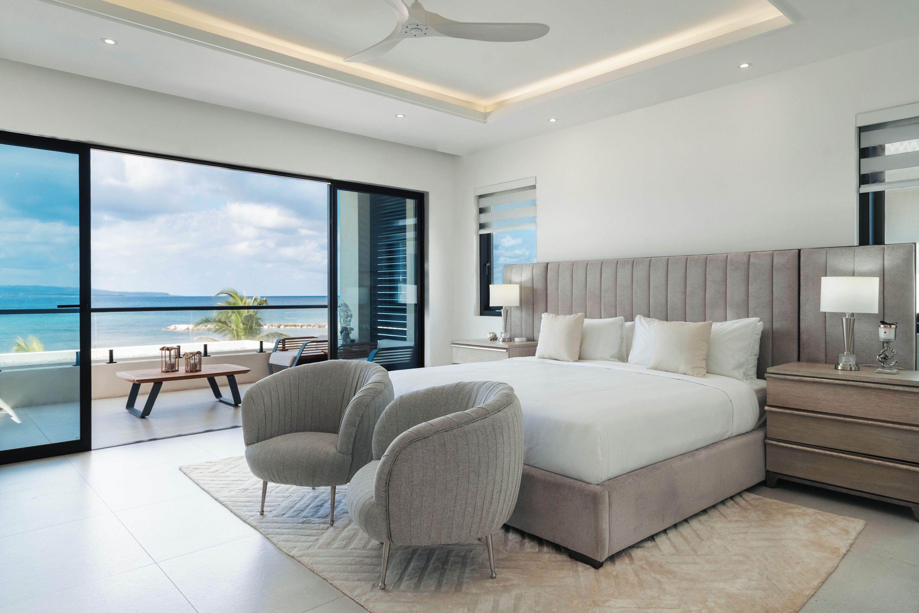 A modern bedroom with a large bed, two chairs, a nightstand, and a view of the ocean.