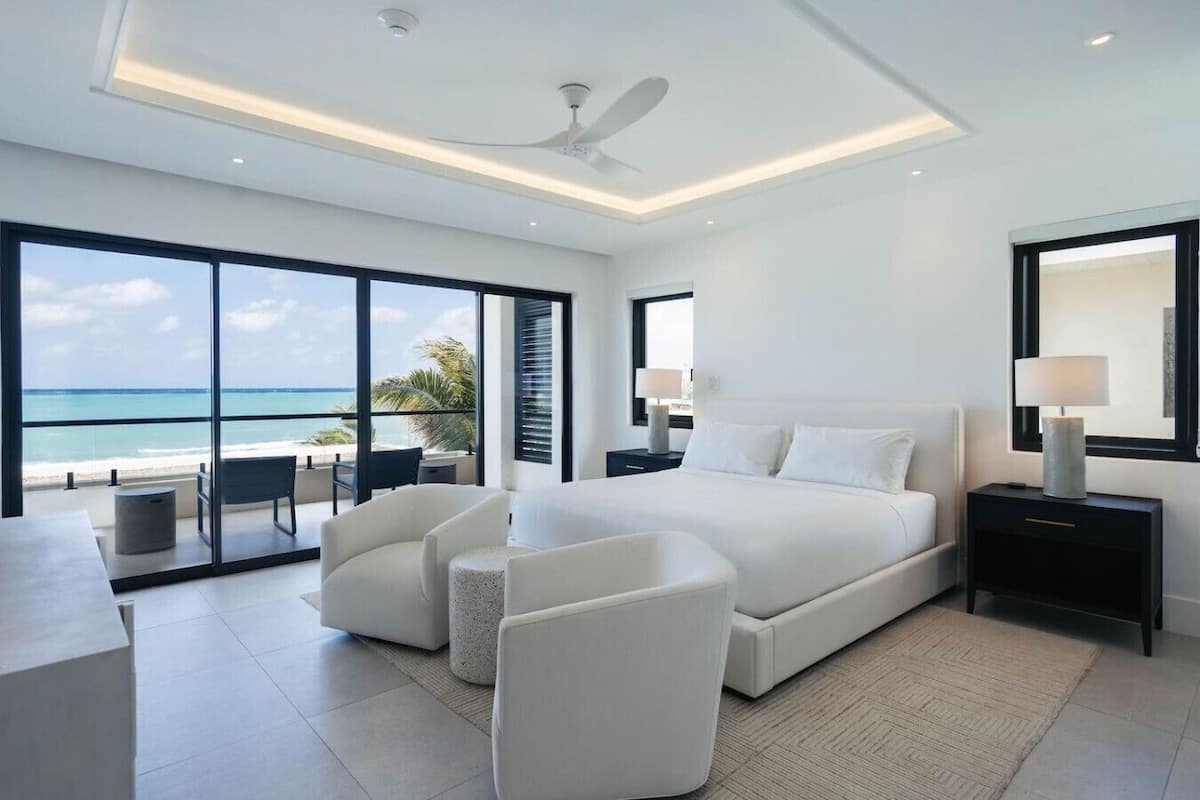 Habitación de hotel moderna con una cama grande, dos sillones blancos, un escritorio y vista al océano.