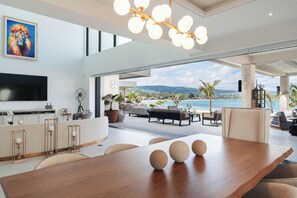 A spacious living area with a large wooden table, a TV mounted on the wall, and a view of the sea. - Bocobay Palm Beach Villas (St. Anns Bay)