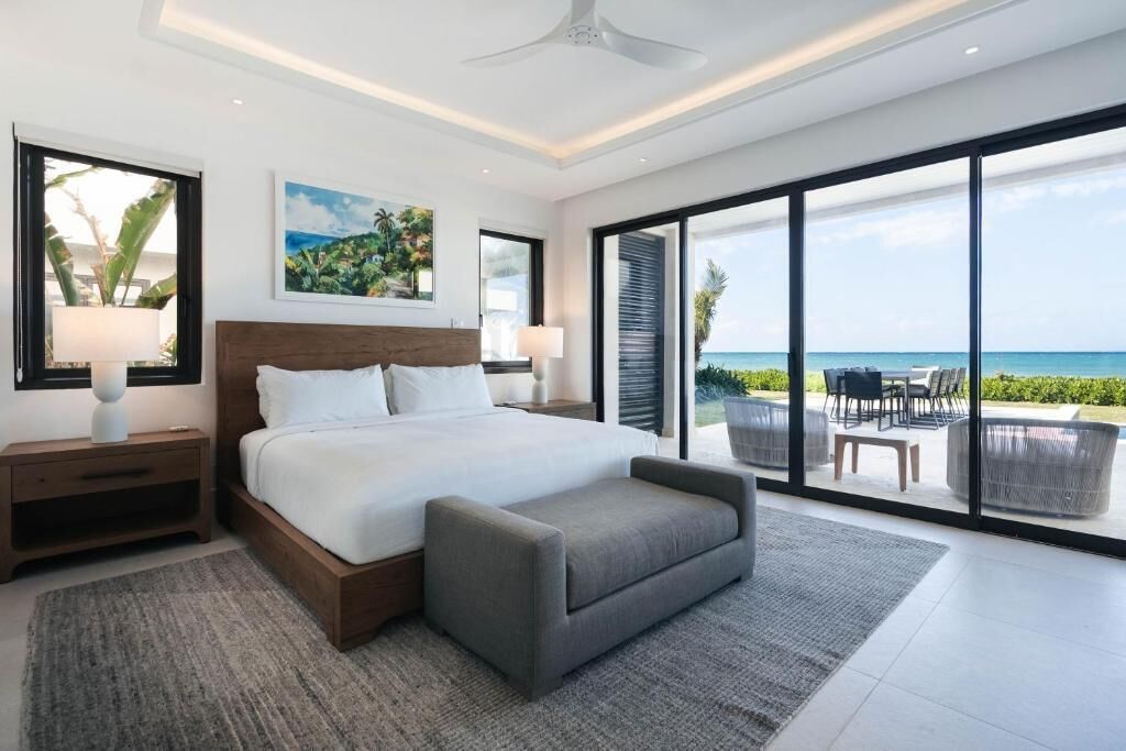 A modern bedroom with a large bed, bedside tables, a grey ottoman, and a view of the ocean through floor-to-ceiling windows.