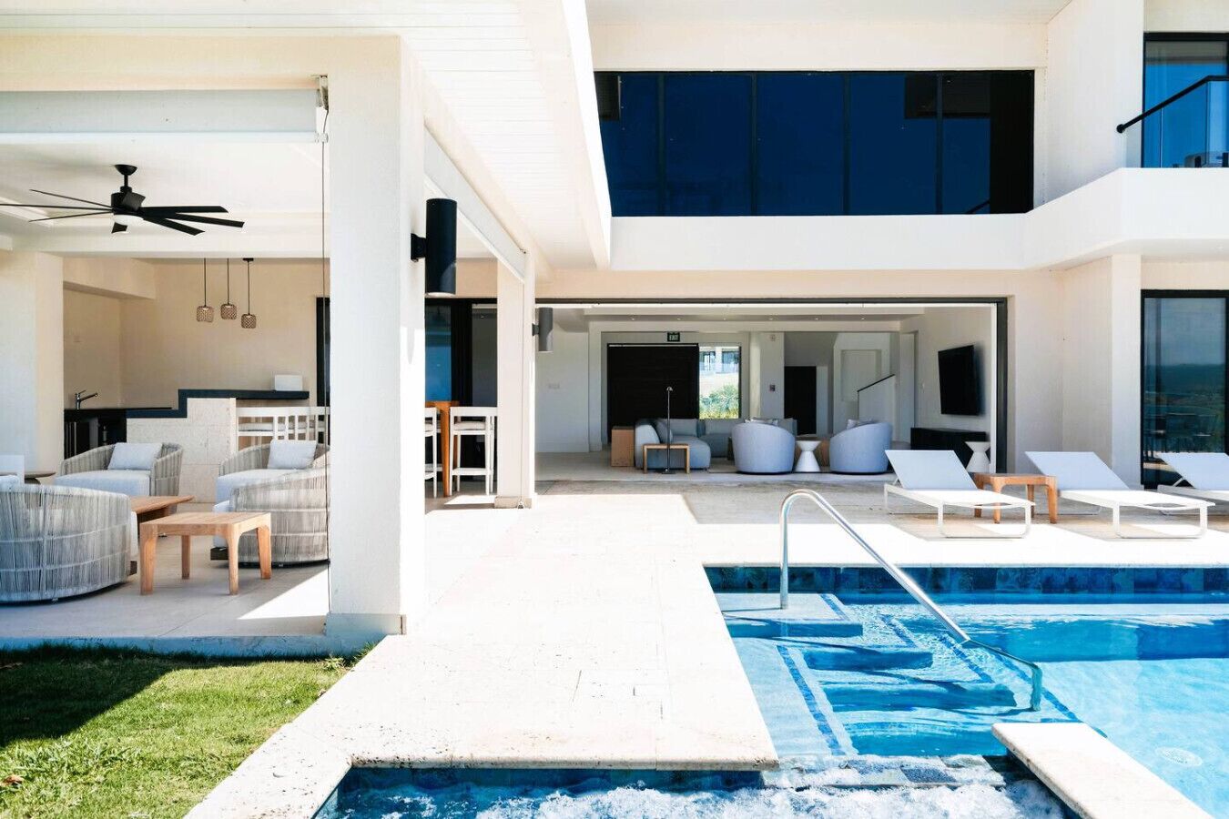 A modern outdoor pool area with lounge chairs, a seating area with sofas, and a kitchen visible through an open door.
