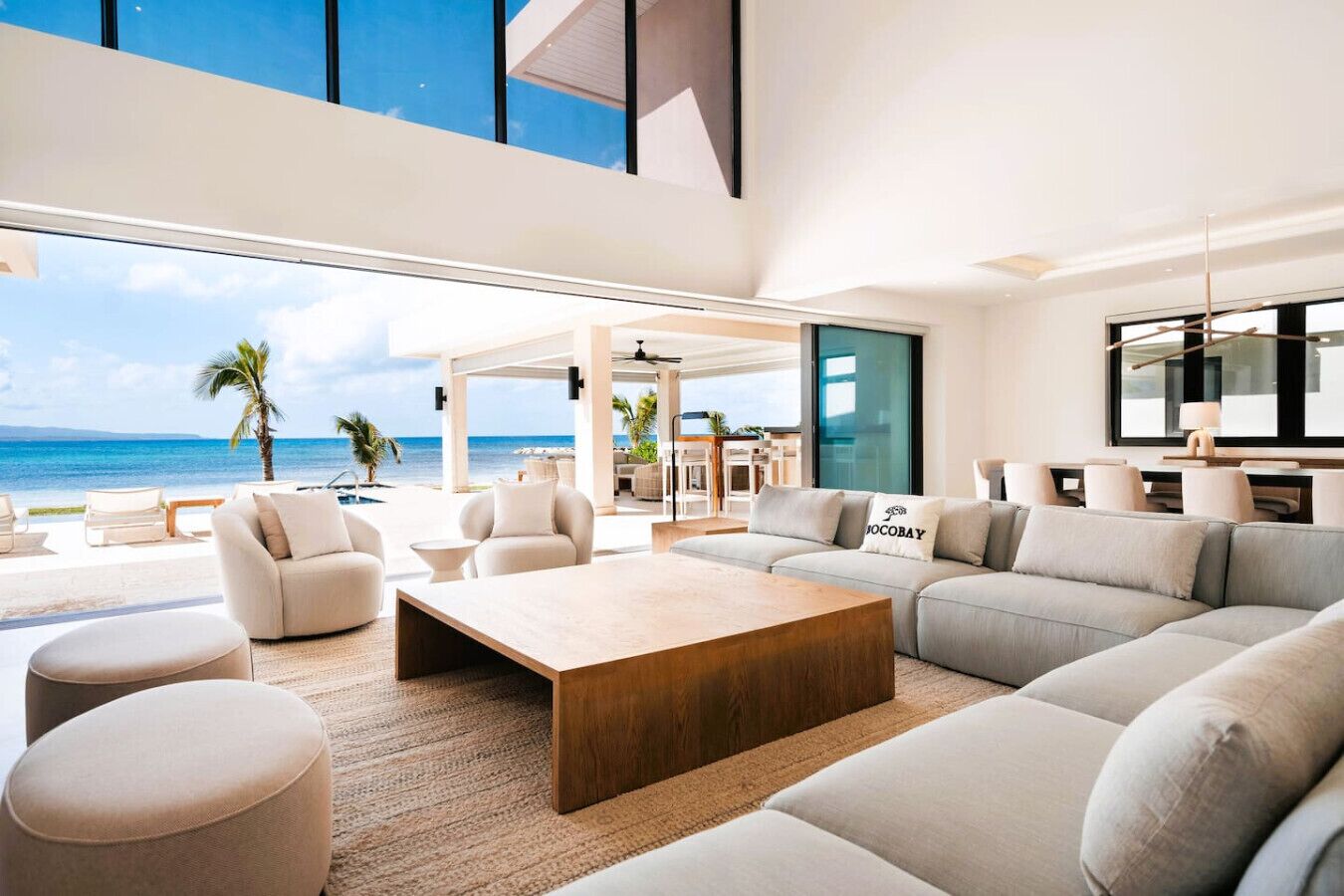 A spacious living area with a large sofa, ottomans, and a coffee table, overlooking a beach and pool.