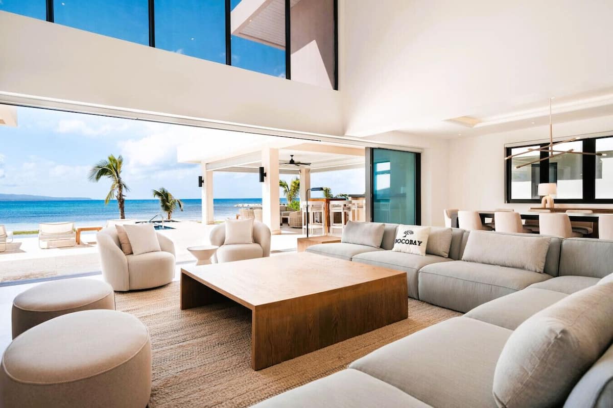 Amplia sala de estar con un sofá grande, otomanas y una mesa de centro, con vistas a una playa y una piscina.