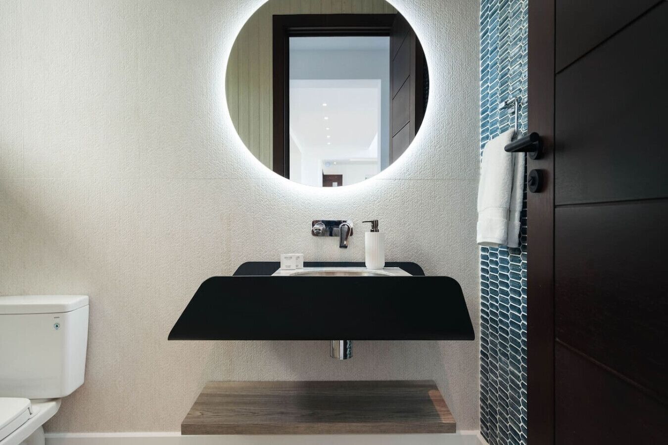 A bathroom with a round mirror, a black sink, a wooden shelf, and a toilet.