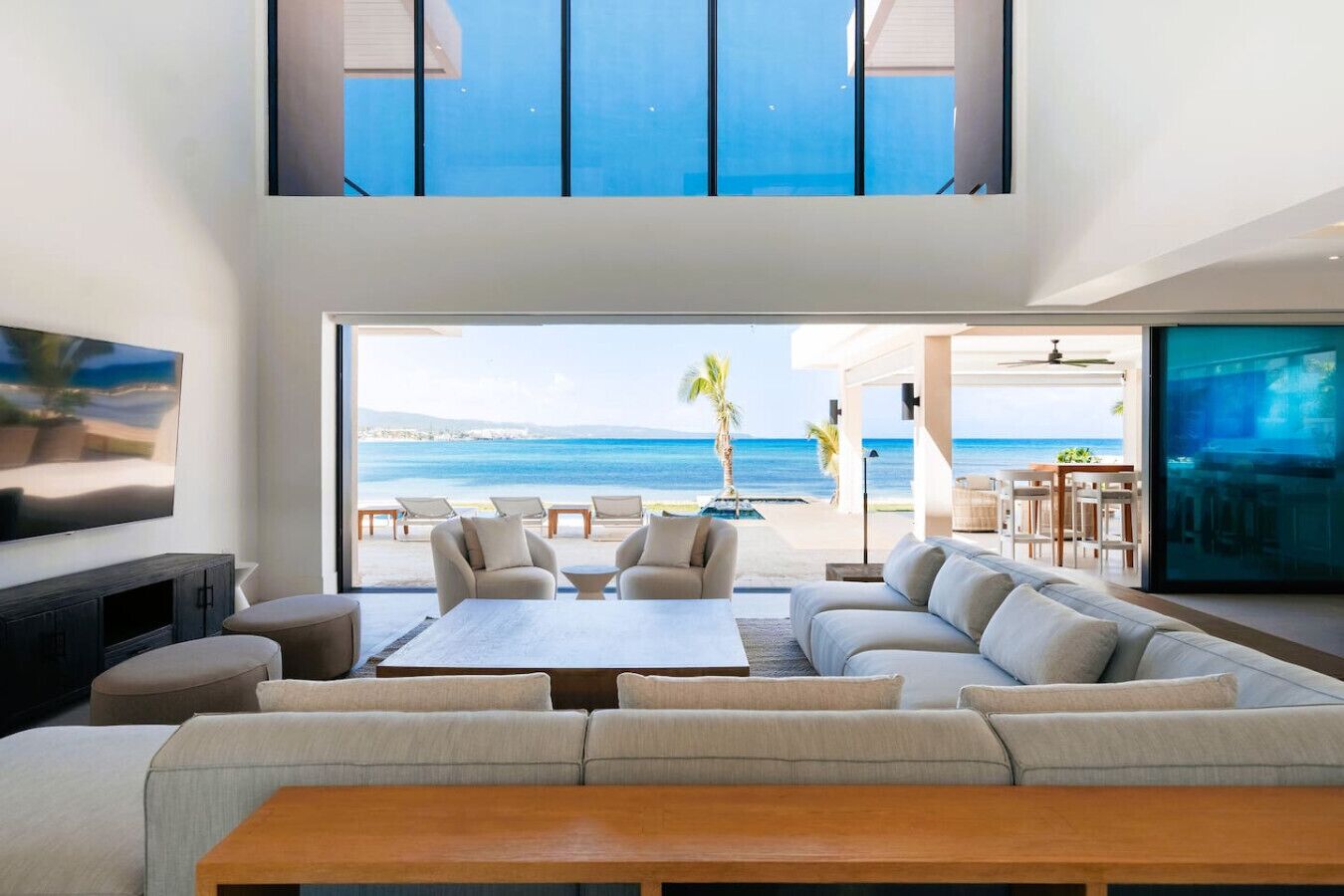 Una sala moderna con un sofá grande, una mesa de centro y una televisión. Hay un balcón con vistas al océano y un comedor con sillas y una mesa.