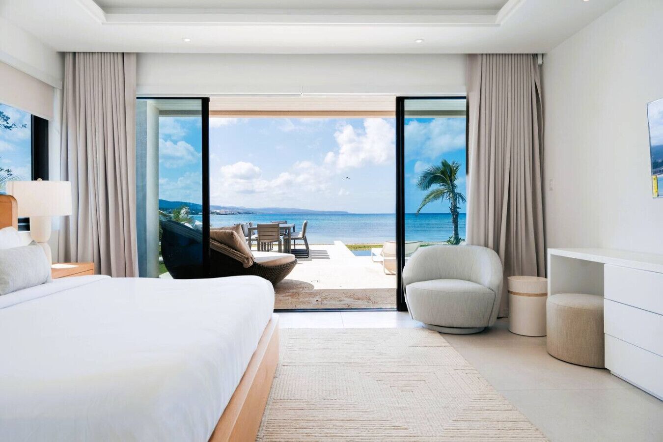A modern hotel room with a large bed, a seating area with a view of the ocean, and a balcony with curtains.