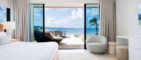 A modern hotel room with a large bed, a seating area with a view of the ocean, and a balcony with curtains.