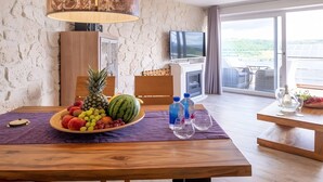 A room with a stone wall, a wooden table with a fruit bowl, a television, and a view of a green landscape through large windows. - Apartment Seehöhe at Maarberg Resort – Holiday Apartment for 4 Guests (48 m²) in Schalkenmehren (Schalkenmehren)