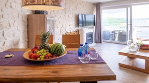 Ein Zimmer mit Steinwand, einem Holztisch mit Obstschale, einem Fernseher und Blick auf eine grüne Landschaft durch große Fenster.