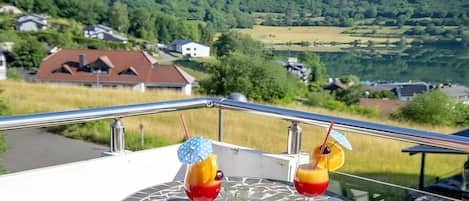 A balcony with two drinks, a table, and a scenic view of houses and hills.