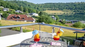 Un balcón con dos bebidas, una mesa y una vista panorámica de casas y colinas.