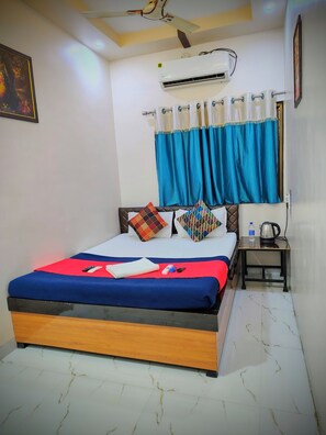 A neatly made bed with a red and blue bedspread, a wooden headboard, and a nightstand with a kettle and water bottle. - Hotel Sai Nanda Shirdi (Shirdi)