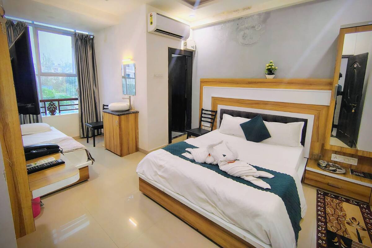 A hotel room with a bed, bedside table, air conditioning unit, window with curtains, and a small potted plant.