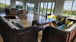 Un salón espacioso con un amplio sofá, una mesa de centro y una mesa de comedor. Varias ventanas ofrecen luz natural y vistas al exterior.