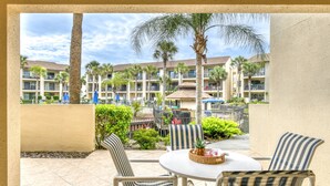 A patio with two striped chairs and a table, overlooking a courtyard with palm trees and a building with balconies. - First Floor Condo, Two Pools, One Heated Pool, Easy Beach Access, Large Patio, $300 in Free Tickets! (St. Augustine Beach)