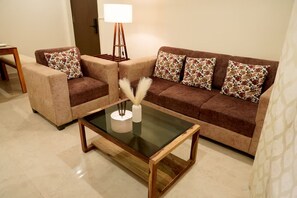 Una sala de estar con un sofá marrón, dos sillones, una mesa de centro de vidrio y una lámpara de pie.