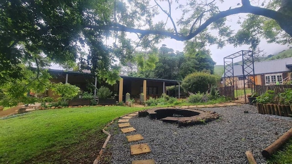 A garden with a circular stone fire pit, a wooden deck, and a metal structure.