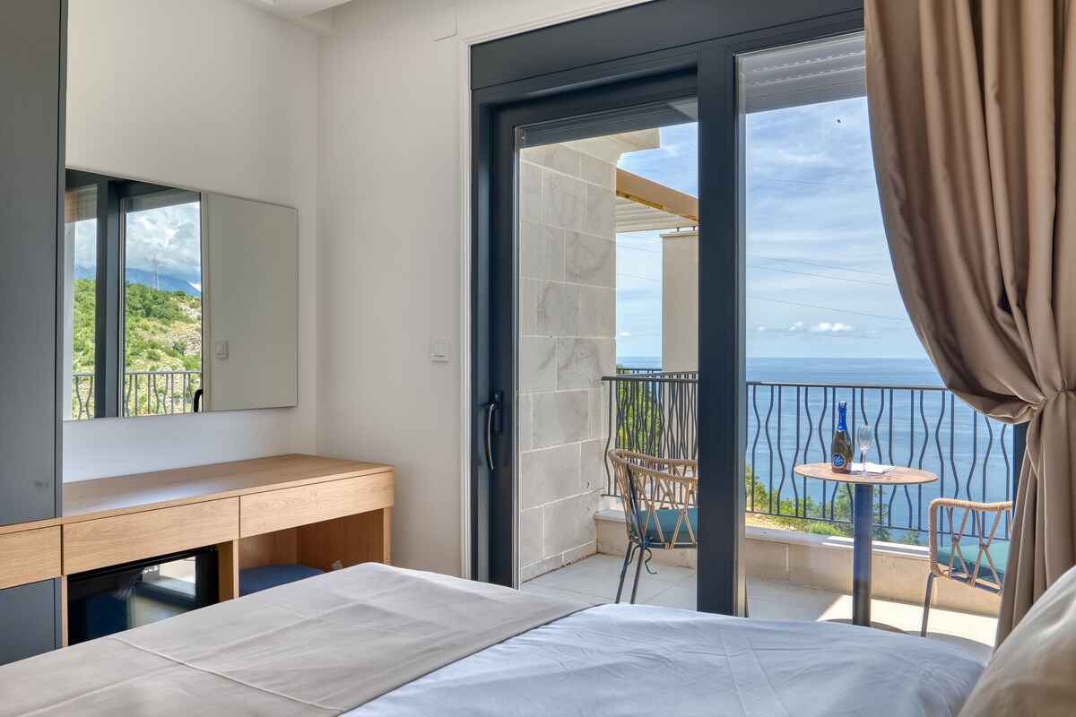 Quarto com cama, escrivaninha de madeira, banheiro e varanda com vista para o mar.