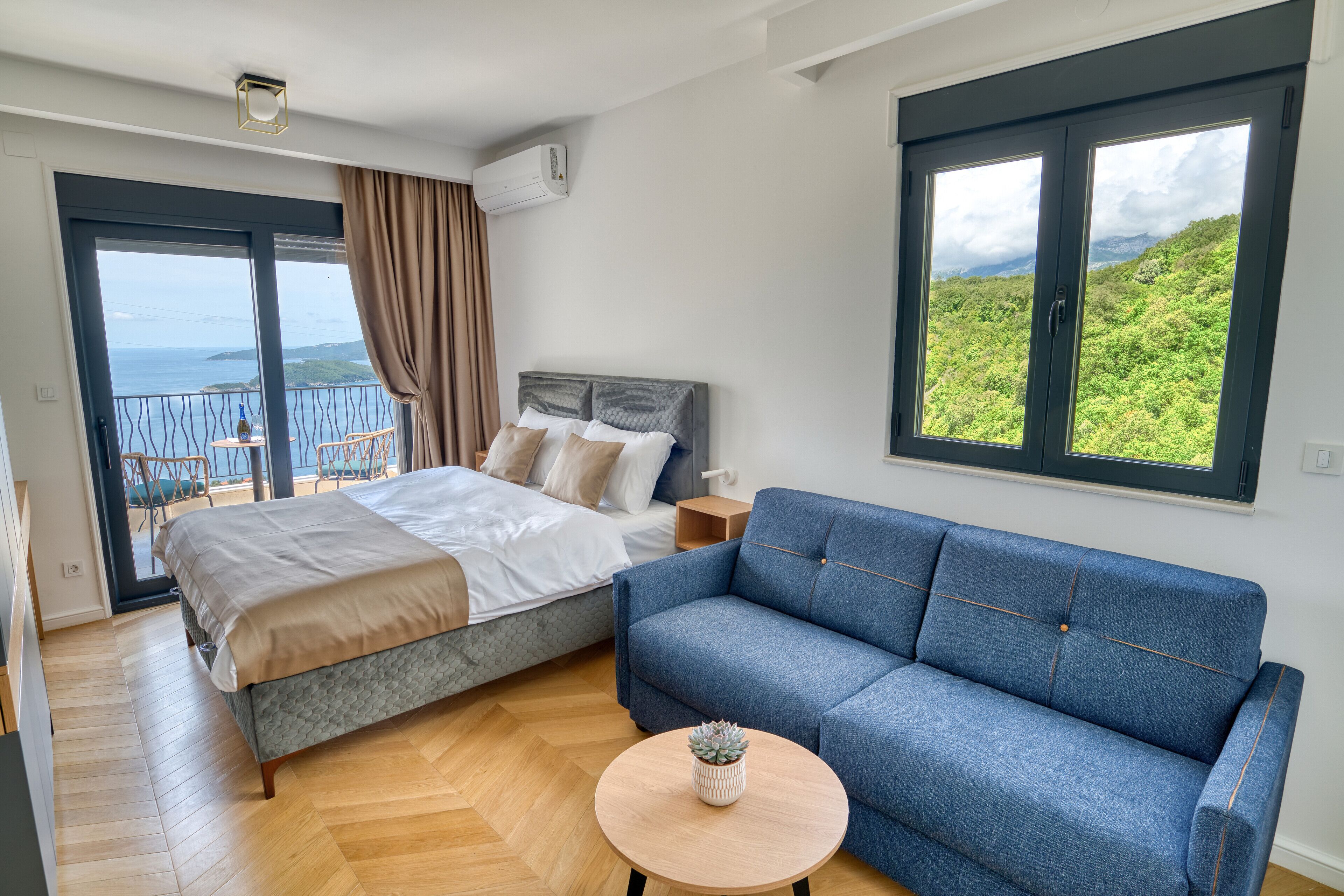 Une chambre avec un lit, un canapé bleu, une petite table ronde avec une plante, un balcon avec vue sur la mer et une fenêtre donnant sur la verdure.