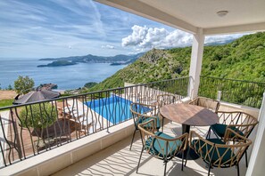 Un balcó amb piscina, cadires de vimet i una taula amb vista panoràmica al mar i les colines.