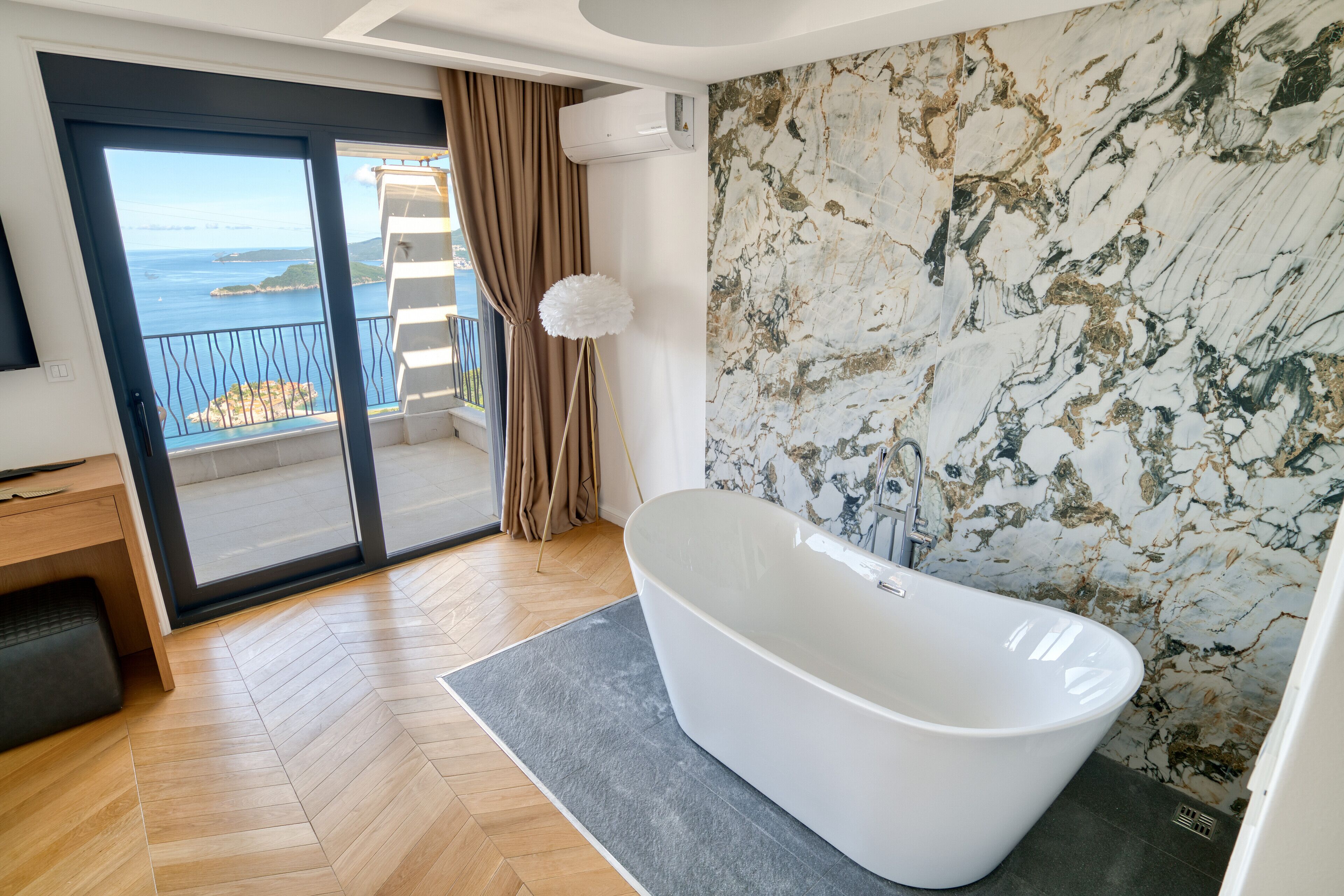 Een moderne badkamer met een vrijstaand bad, een houten vloer met een visgraatmotief, een marmeren wand en een balkon met uitzicht op zee.