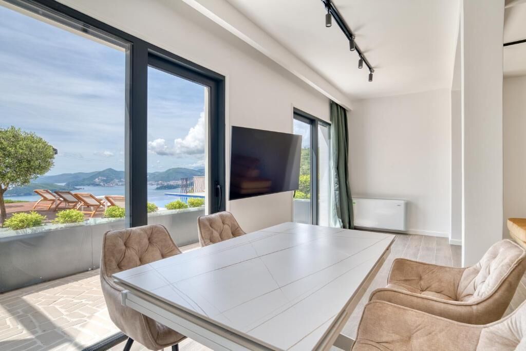 A modern room with a large table, comfortable seating, a flat-screen TV, and a balcony with a view of the mountains.