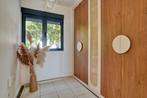 Una habitació amb finestra, porta de fusta, mirall rodó i un vas amb plantes seques.