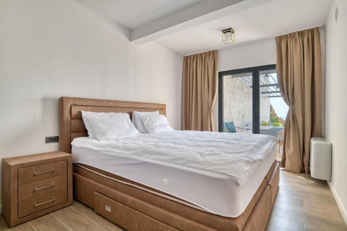 Quarto com cama, gavetas de cabeceira, janela com cortinas e vista para o exterior.