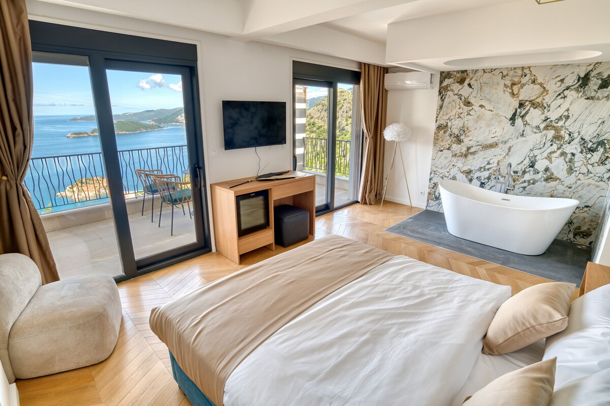 Quarto de hotel moderno com uma cama grande, televisão, secretária, banheira e varanda com vista para o mar.