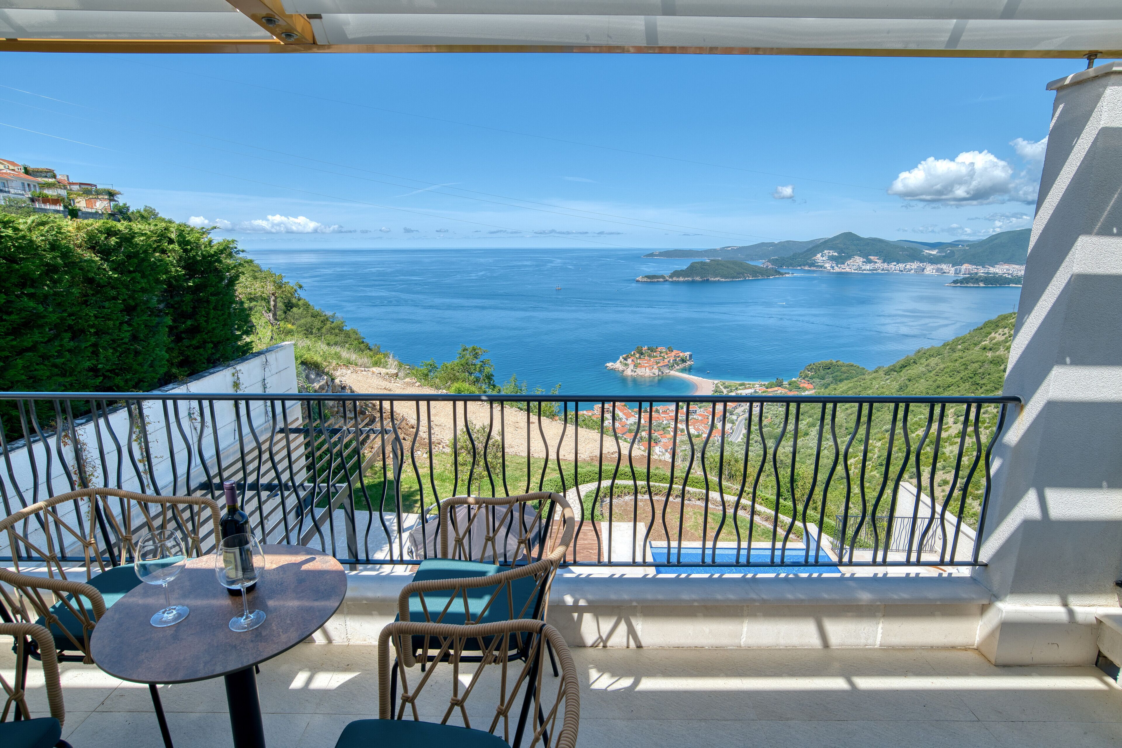 A balcony with wicker chairs and a table, overlooking a coastal view with a swimming pool and buildings in the distance.