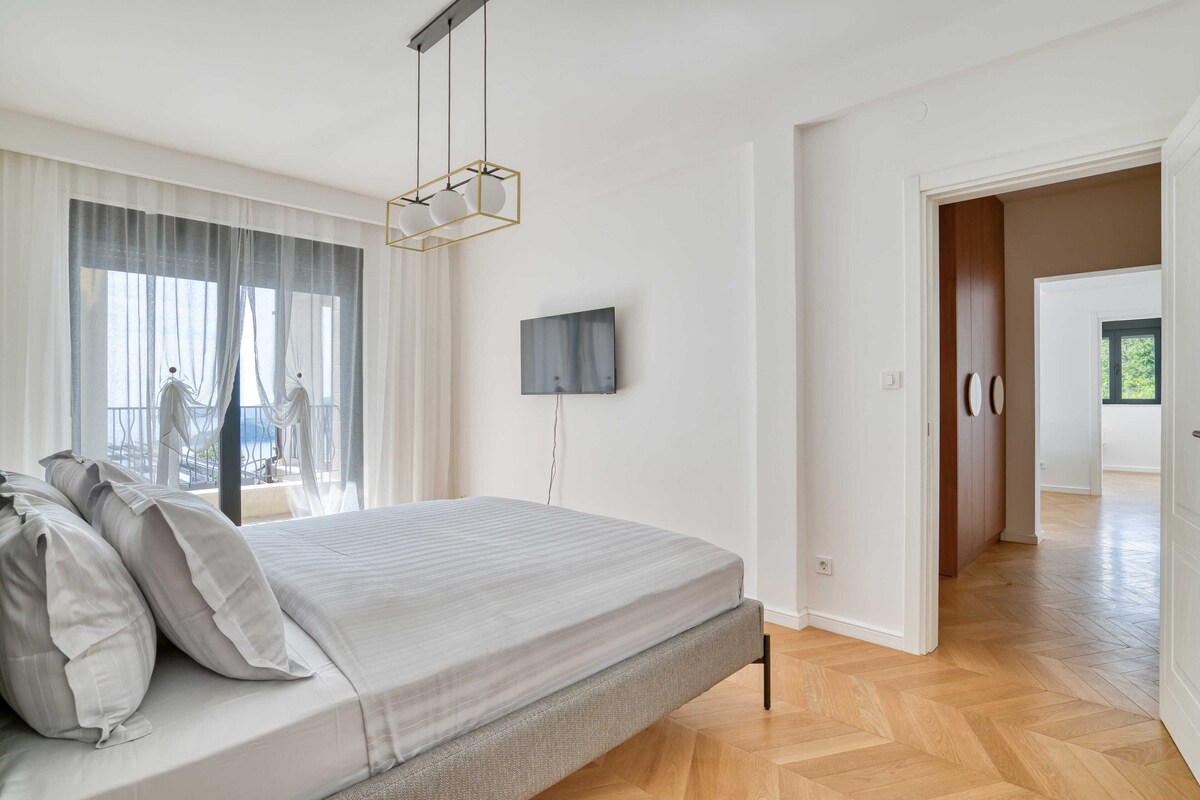 Quarto moderno com cama, televisão fixada na parede e vista do exterior através de grandes janelas.