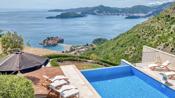 Une piscine avec des chaises longues, une terrasse en bois et une vue sur la mer et les collines.