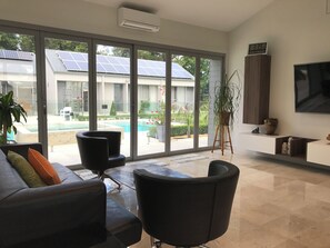 Una sala de estar moderna con un amplio ventanal corredera que da a una zona de piscina, un televisor de pantalla plana montado en la pared y una planta en maceta.