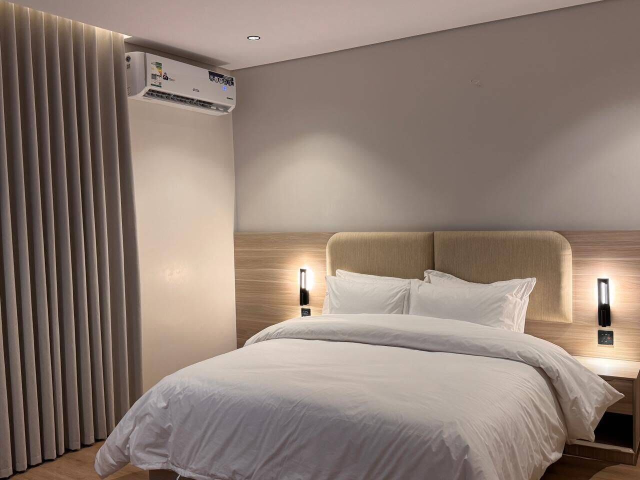 Una camera d'hotel moderna con un letto grande, due comodini con lampade a parete, un condizionatore d'aria verticale e un'ampia finestra con tende.