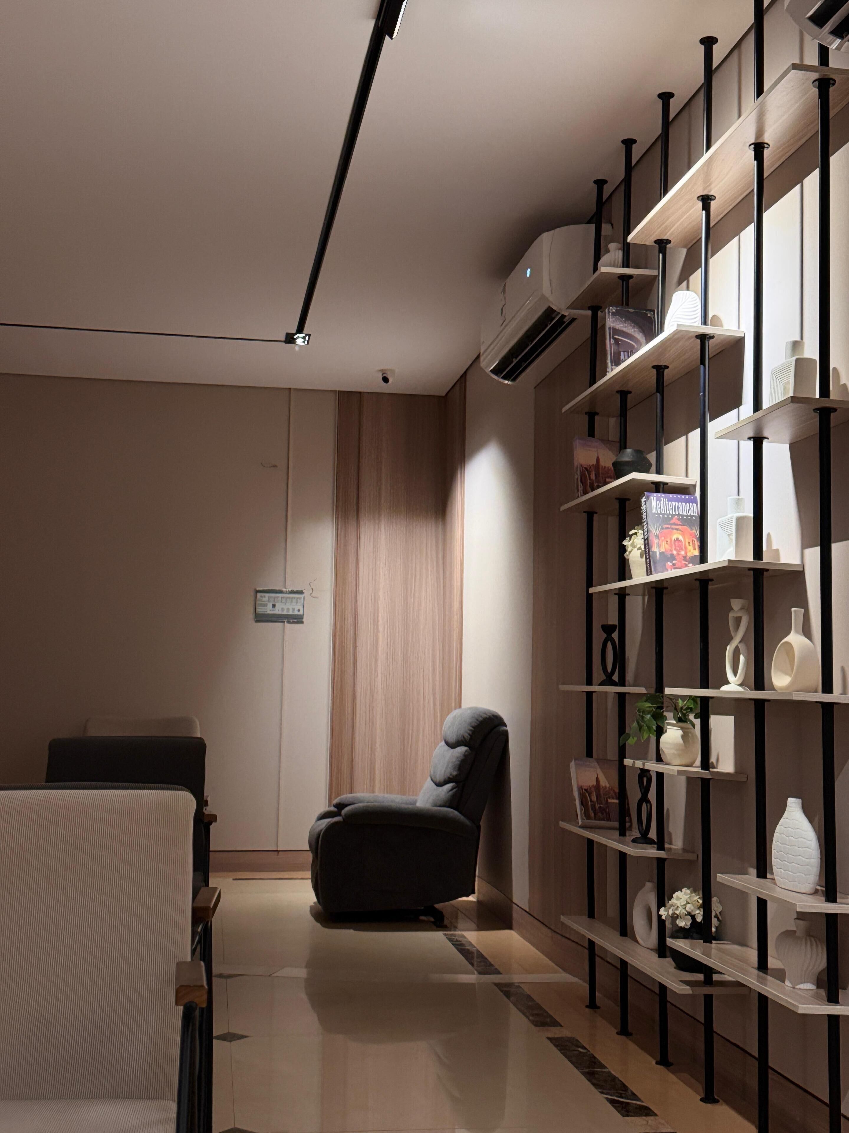 A modern room with a dark armchair, a wooden panel wall, a bookshelf with decorative items, and a ceiling-mounted air conditioner.