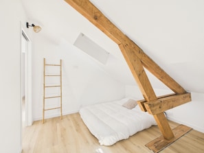 Ein Zimmer mit schrägen Wänden, einer Holzleiter, einem Bett und einem kleinen Fenster.