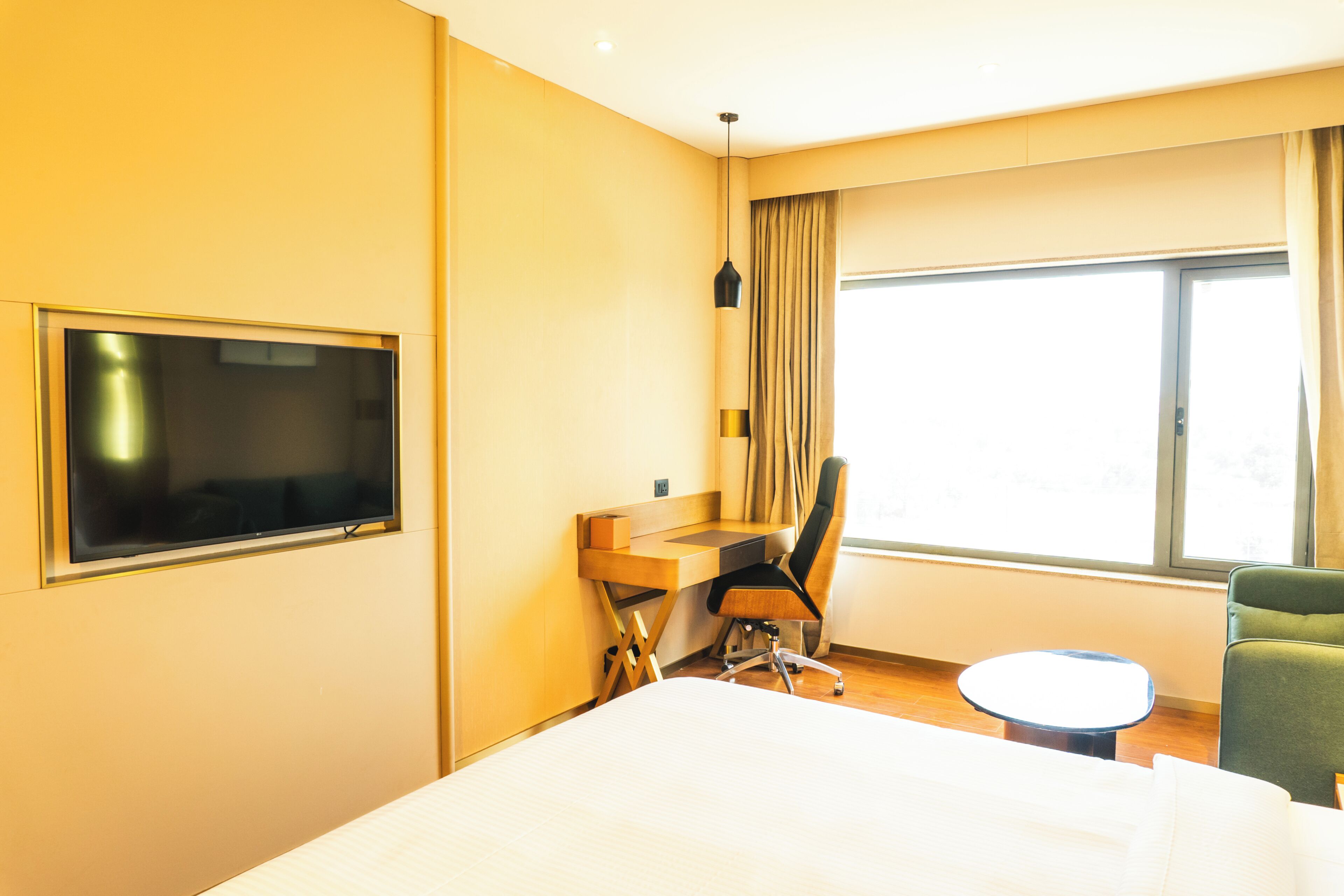 A hotel room with a bed, a desk with a chair, a television, a window with curtains, and a small round table.