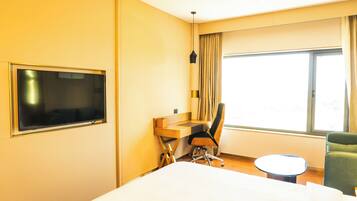 A hotel room with a bed, a desk with a chair, a television, a window with curtains, and a small round table.
