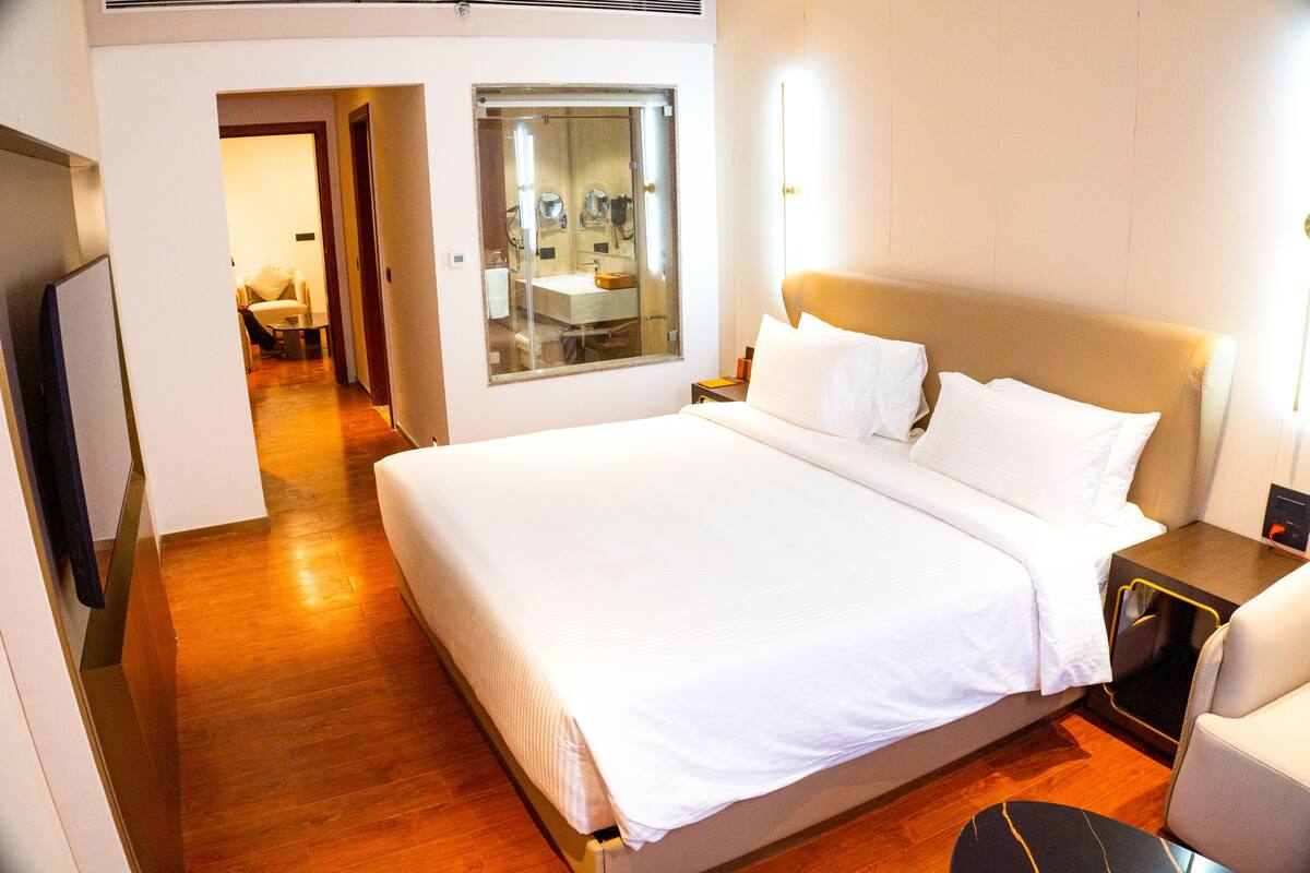 Habitación de hotel con una cama grande, una mesita de noche, una silla, un espejo y un baño visible a través de una puerta de vidrio.