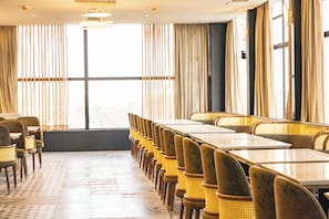 A restaurant with a long table and chairs, large windows with curtains, and a tiled floor.