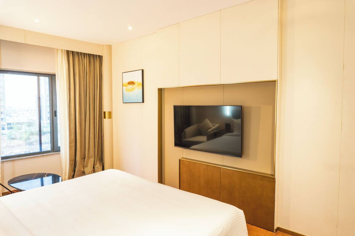 Habitación de hotel con cama, televisor montado en la pared, ventana con cortinas y una mesita.