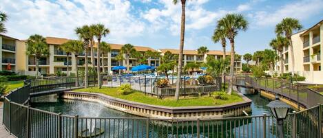 A resort with a central water feature, surrounded by palm trees and a pool area with umbrellas.