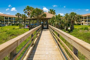 A wooden boardwalk leading to a building with a thatched roof, surrounded by palm trees and a clear blue sky. - First Floor Condo, Two Pools, Large Patio, Outdoor Dining, Direct Beach Access, $300 in Free Tickets (St. Augustine Beach)