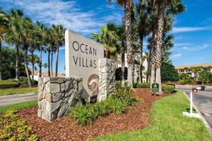 A sign for Ocean Villas with a stone base, palm trees, and a clear sky. - First Floor Condo, Two Pools, Large Patio, Outdoor Dining, Direct Beach Access, $300 in Free Tickets (St. Augustine Beach)