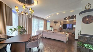 Ein modernes Wohnzimmer mit einem Ecksofa, einem Esstisch, einer Pendelleuchte und einem Flachbildfernseher.
