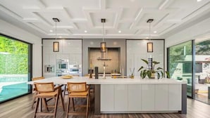 A modern kitchen with a central island, wooden chairs, pendant lights, and a view of a swimming pool. - Willow Glen (Los Angeles)