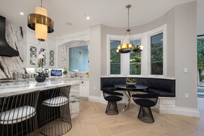 A modern kitchen with a marble countertop, black bar stools, a black upholstered bench, and a large window with a view of greenery. - Gardner Modern (Los Angeles)