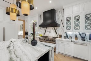 A modern kitchen with white cabinetry, a marble countertop, a black range hood, and pendant lighting. - Gardner Modern (Los Angeles)