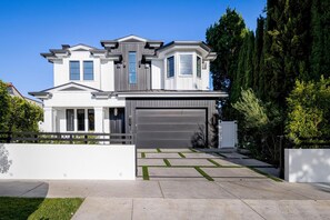 A modern two-story house with a garage, a well-maintained lawn, and tall trees in the background. - Gardner Modern (Los Angeles)